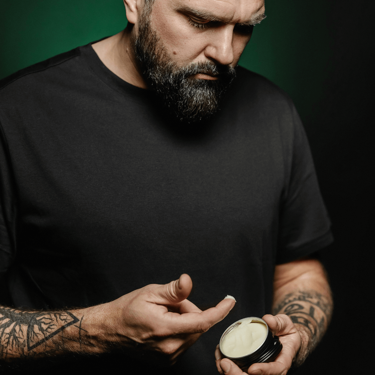 Mand med fuldskæg og tatoverede arme i sort t-shirt tager skægplejeprodukt fra sort krukke med fingeren, klar til at style og pleje skægget.