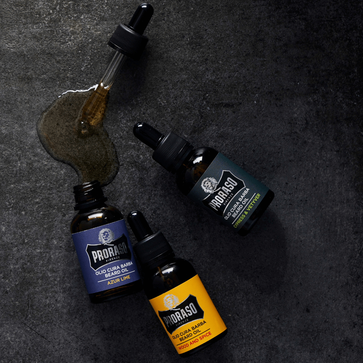 Tre flasker Proraso skægolie med pipette i varianterne Azur Lime, Cypress & Vetyver samt Wood and Spice, placeret på mørk baggrund med spildt olie for et stilrent udtryk.